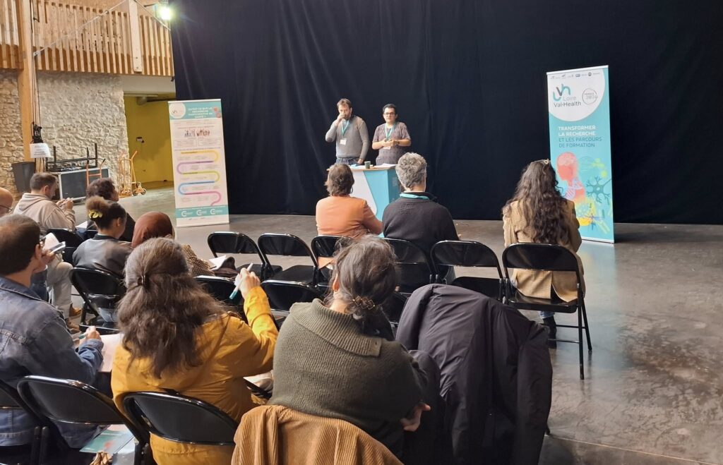 Des participants assis regardent Thomas et moi restituer notre atelier. Nous sommes debout derrière un pupitre aux couleurs du projet Loire Val Health (bleu et blanc) et Thomas boit son café pendant que j'explique ma partie. Les participants nous écoutent attentivement.
